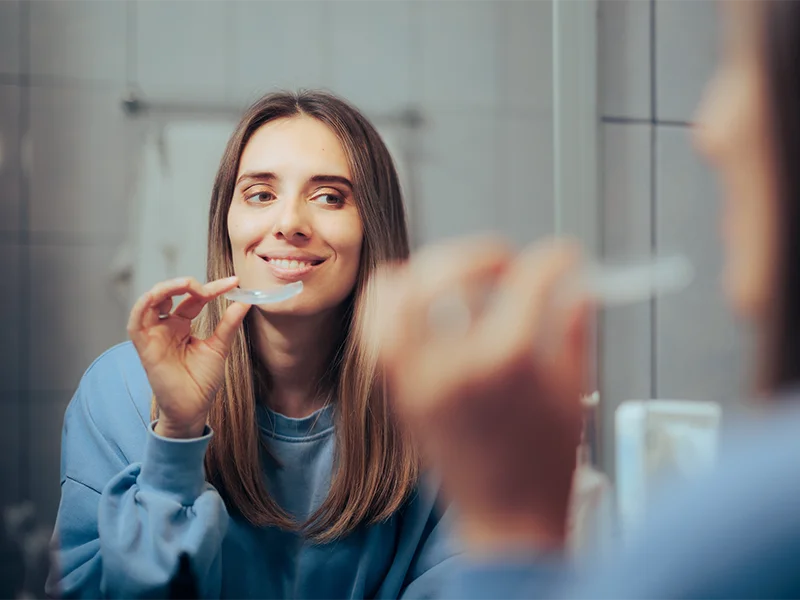 Woman holding a clear dental aligner while looking in the bathroom mirror before inserting it.